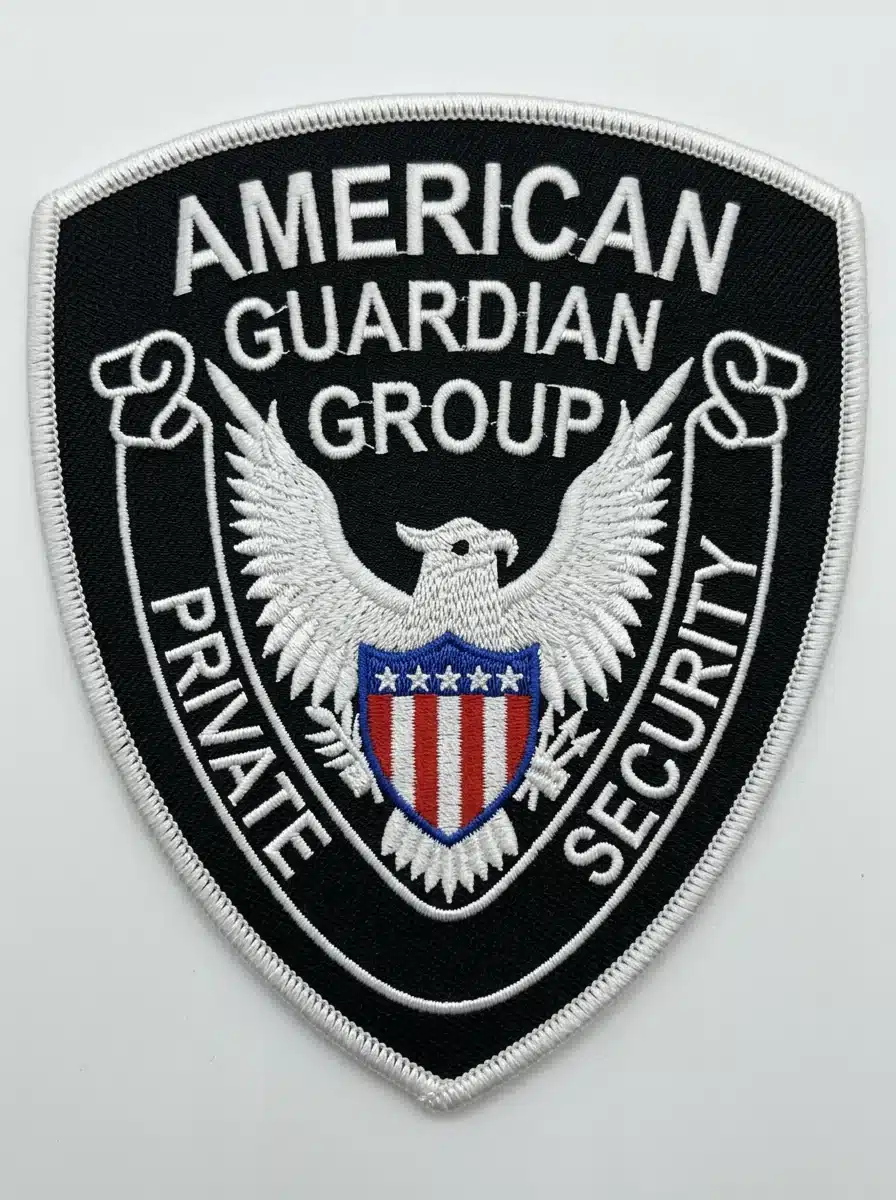 Embroidered shield security patch in black with white lettering, a white spread-wing eagle holding a red, white and blue shield, a scroll banner and a white merrowed border