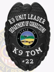 Featuring a polished custom metal badge in black color, this design includes a shield shape with "K9 UNIT LEADER," "DEPARTMENT OF CORRECTIONS," and "K9 TOM" clearly inscribed around a central emblem representing Ohio, along with the number "#22." The badge is completed with a safety pin attachment for easy application on uniforms.