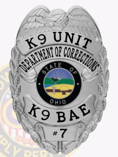 An eye-catching custom metal badge in silver color, designed as a shield with "K9 UNIT" and "DEPARTMENT OF CORRECTIONS" prominently displayed at the top. The central design illustrates the state seal of Ohio, complemented by "K9 BAE" and "#7," equipped with a safety pin attachment for secure wear.