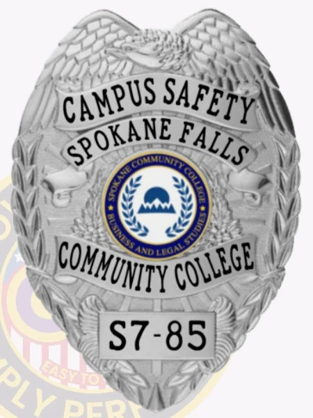 A sleek custom metal badge in silver color with a safety pin attachment, prominently displaying "CAMPUS SAFETY SPOKANE FALLS" at the top. The badge features a central emblem for Spokane Community College, highlighting its focus on business and legal studies, with the identifier "S7-85" at the bottom for clear identification.