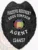 A striking image of a custom metal badge in black color, featuring a detailed shield design adorned with the words “Fugitive Recovery” and the name “Doug Simpson,” complete with a central Alabama seal and a safety pin attachment for secure wear.