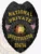 A striking custom metal badge in black color with a safety pin attachment, prominently featuring "NATIONAL PRIVATE INVESTIGATOR" in bold yellow lettering. The badge includes the Alabama state seal and small flags, emphasizing its professional design and authority.
