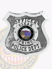 Silver metal shield badge custom-made for Clear Springs police officer, displaying black text "OFFICER" at the top, "CLEAR SPRINGS" encircling a vibrant State of Alaska seal in the center, and "POLICE DEPT." at the bottom, attached with a safety pin.