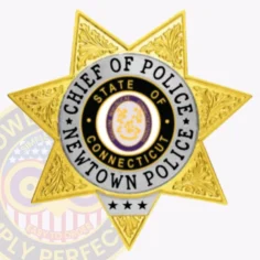 Metal badge in gold shaped as a 7 point star for newtown police the chief of police, with embossed decorative leaves on each point, a round emblem in the center showing the connecticut state seal, and a white circular band containing black text that reads “chief of police newtown police,” fastened by a safety pin.