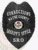 A custom metal badge in a sleek black color, shaped like a shield, representing the Wayne County Sheriff's Office for Corrections. The badge prominently displays "CORRECTIONS" at the top and "WAYNE COUNTY" below it in bold white letters. In the center, it features the state seal of Arkansas, surrounded by decorative elements. The badge also reads "SHERIFF'S OFFICE" and "SRO" at the bottom in white text, and includes a safety pin attachment on the back, allowing for secure wear by school resource officers, emphasizing its functional design and authority.