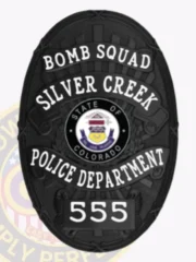 Oval-shaped custom black metal badge for Silver Creek Police Bomb Squad showing the Colorado state seal in the middle, surrounded by white lettering "BOMB SQUAD," "SILVER CREEK," "POLICE DEPARTMENT," and badge number "555," held by a safety pin.