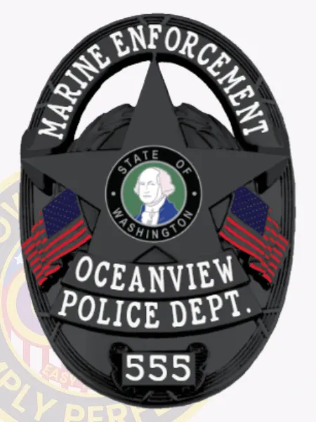 Oval-shaped custom black metal badge for Oceanview police Marine Enforcement highlighting a five-point star at its core with the State of Washington emblem, clear white text MARINE ENFORCEMENT at the top edge, OCEANVIEW POLICE DEPT. below, badge number 555 displayed on a scroll underneath, flanked by two U.S. flags, designed with a safety pin attachment.