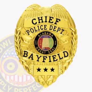 A detailed view of a custom metal badge in gold color featuring an eagle design at the top. The badge prominently displays the title "CHIEF" and includes the text "POLICE DEPT. BAYFIELD" along with the Alabama state seal in the center. The badge number is marked with stars at the bottom, and it is designed with a safety pin attachment for secure fastening.