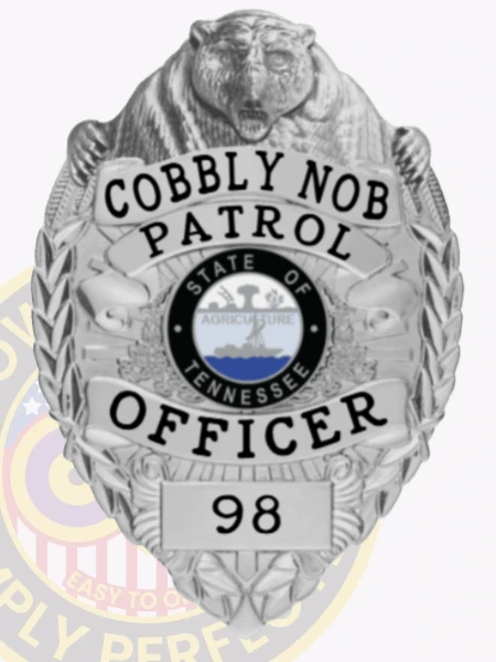 A detailed view of a custom silver badge with safety pin attachment, presenting "Cobbly Nob Patrol" and "OFFICER" in bold letters. The badge features a bear motif at the top and intricate designs, emphasizing its official purpose and elegant appearance.