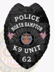 Alt Text: A bold custom metal badge all black with white lettering, shaped like a shield and featuring intricate textures. The badge displays "POLICE" at the top and "NORTH HAMPTON" beneath it in prominent white letters. In the center, the state seal of Utah is shown within a circular design, flanked by two small American flags. Below the seal, "K9 UNIT" is inscribed in white, and the number "62" is displayed at the bottom on a black panel. The matte black finish of this custom metal badge all black with white lettering enhances its professional and authoritative presence, representing the dedication of K9 officers in law enforcement.