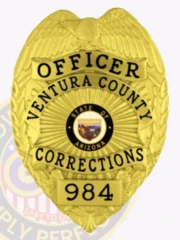 A custom metal badge in a bright gold color, shaped like a traditional shield, representing the Ventura County Corrections. The badge features "OFFICER" at the top, followed by "VENTURA COUNTY," and "CORRECTIONS" prominently displayed. In the center, it showcases the state seal of Arizona, framed by detailed designs. The badge is numbered "984" at the bottom and includes a safety pin attachment on the back, allowing for secure wear by correctional officers, emphasizing its practical application and distinctive design.
