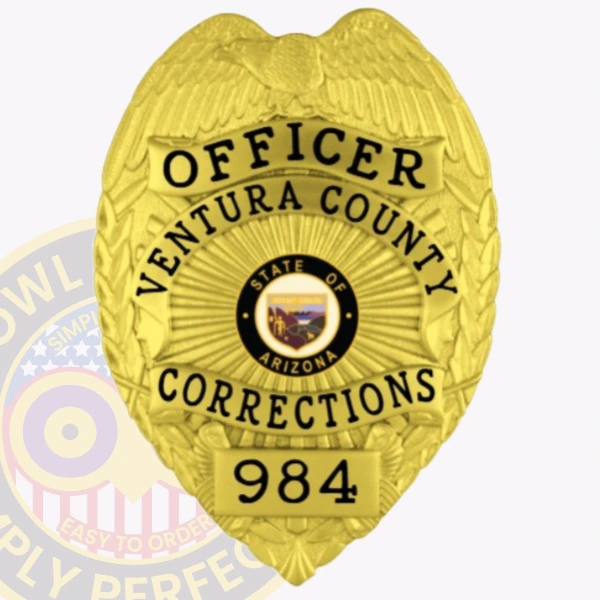 A close-up of a custom metal badge in a brilliant gold color, shaped like a shield, representing the Ventura County Corrections. The badge displays "OFFICER" at the top, followed by "VENTURA COUNTY" and "CORRECTIONS" in bold text. In the center, it features the state seal of Arizona, surrounded by decorative patterns. The badge is marked with the number "984" at the bottom and includes a safety pin attachment on the back, allowing for secure attachment to uniforms, showcasing its practical design for correctional officers.