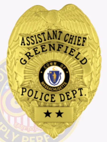 Gold metal badge for Greenfield Police Assistant Chief with an eagle emblem at the top, black engraved text reading "ASSISTANT CHIEF" and "GREENFIELD POLICE DEPT.," centered Massachusetts state seal, two black stars on a plate below, and safety pin fastener.
