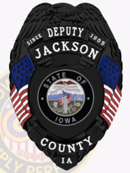 A custom metal badge in black featuring the title "DEPUTY" at the top, along with the name "JACKSON" and "SINCE 1905." The center showcases a circular emblem of the state of Iowa, flanked by decorative blue panels and American flags on either side. Below the emblem, the badge reads "COUNTY IA." The matte black finish gives the badge a sleek and modern appearance, emphasizing its official status and authority in law enforcement.