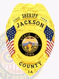 A custom metal badge in gold displaying the title "sheriff" at the top, with the name "jackson" and "since 1905" above it. The center features a circular emblem of the state of iowa, surrounded by decorative blue panels and american flags on either side. Below the emblem, the badge reads "county ia. " the badge has a polished gold finish that highlights its official status and intricate design, emphasizing its significance as a symbol of authority in law enforcement.