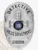 Silver oval custom metal badge for Rivertown police detective with safety pin back, displaying embossed urban buildings on a radiant silver background, black text banners with "DETECTIVE," "POLICE DEPARTMENT," and "RIVERTOWN," featuring the New York state seal in the middle.