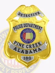 Custom gold eagle top shield metal badge for Pine Creek police detective showing a large gold eagle on top, gold textured shield body with silver text panels labeled "DETECTIVE," "POLICE DEPARTMENT," "PINE CREEK ALABAMA," and the Alabama Great Seal in the center, fixed with a safety pin on the back.