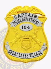Custom gold metal badge shaped as a shield with eagle figure on top, silver panels with black lettering "CAPTAIN" and "POLICE DEPARTMENT," a central silver oval marked "184," and a silver bottom banner labeled "GREAT LAKES VILLAGE," secured by safety pin.