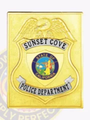 Square gold metal badge for Sunset Cove police officers, featuring an embossed eagle perched above the central shield displaying the colorful State of California seal, framed by silver ribbons reading "Sunset Cove" and "Police Department" in black letters; attached with safety pin.