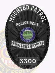 An elegant custom metal badge in black with safety pin attachment, showcasing "BROOKSHIRE HEIGHTS" prominently across the center. The design includes the state seal of Vermont and features intricate patterns along the edges. The matte finish adds a sleek touch, making it suitable for law enforcement use.