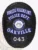 A distinctive custom metal badge in black with reverse enamel blue accents and with safety pin attachment displays "DOMESTIC VIOLENCE UNIT" prominently at the top in large white text. Below it, "POLICE DEPT." stands out clearly, while "OAKVILLE" appears in striking blue. The central design includes the state seal of Georgia, and "043" is elegantly positioned in a yellow box at the bottom.