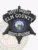 A striking black custom metal badge for deputy sheriffs features the title "DEPUTY SHERIFF ELM COUNTY" in bold lettering along the top. The center displays a round emblem representing the "STATE OF IDAHO," encircled by a blue ring. The bottom inscription reads "SHERIFF'S DEPARTMENT," all secured with a reliable safety pin attachment for easy wear.