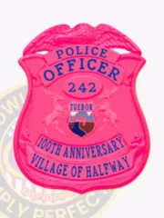 Bright pink metal badge with an eagle emblem crowning the shield, blue text including "POLICE OFFICER," the number "242" inside a central oval, the multicolor Michigan state seal between two deer, and a banner reading "100TH ANNIVERSARY VILLAGE OF HALFWAY," a custom BCAM pink eagle top metal badge with safety pin attachment.