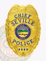 Gold metal badge for Seville Police Department Chief showing a raised eagle atop the shield, blue banners with bold text CHIEF, SEVILLE, and POLICE, a colorful Ohio state seal in the center, and four blue stars near the bottom, designed with a safety pin attachment.