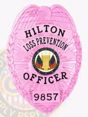 Detailed image of a bright pink metal badge designed for Hilton Loss Prevention Officers, topped with an eagle emblem, black lettering "HILTON LOSS PREVENTION OFFICER 9857," and a central patriotic seal showing stars and American flags, attached with a safety pin.