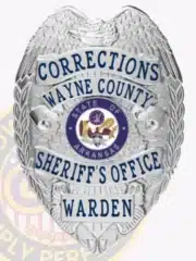Detailed photo of a silver law enforcement badge for Wayne County Corrections, showing a raised eagle design on top, silver ribbons with blue text stating "Corrections Wayne County Sheriff's Office Warden," and the Arkansas state emblem in the center.