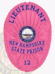 Custom BCAM pink oval metal badge with blue lettering for a Lieutenant at New Hampshire State Prison. The badge displays the New Hampshire state seal prominently in the center and includes a safety pin attachment on the back.