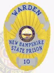 Oval-shaped metal badge in gold with silver panels and blue letters reading "Warden New Hampshire State Prison" along with the number "10." The center displays the official New Hampshire state seal, with a safety pin for secure attachment.