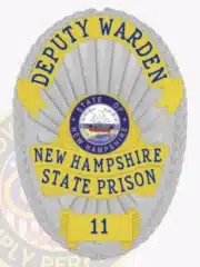 Custom silver oval metal badge with gold panels and blue lettering for the Deputy Warden of New Hampshire State Prison. The badge features the New Hampshire state seal in the center and includes a safety pin attachment on the back.