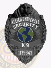 Front view of an Allied Universal custom black metal K9 badge featuring an embossed bear figure at the top, a globe graphic in blue and green center, clear white text stating "Security K9," and a visible safety pin attachment for easy fastening.