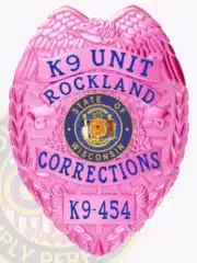 Close-up of a custom BCAM pink metal badge featuring an eagle at the top, labeled "K9 Unit Rockland Corrections," with the State of Wisconsin seal in the center and a safety pin attachment on the back.