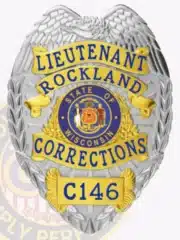 Detailed photo of a silver and gold law enforcement badge for Rockland Corrections, showing an eagle crest on top, gold banners with blue lettering for "Lieutenant Rockland Corrections," the Wisconsin state emblem in the center, and badge number C146 at the bottom.