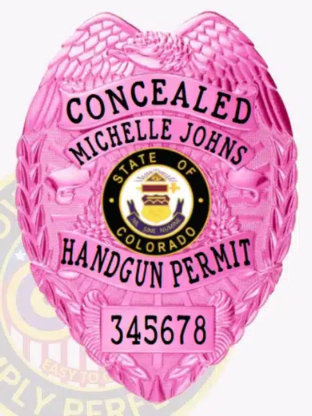 Custom BCAM pink metal badge for concealed handgun permit featuring a detailed eagle design, engraved with "CONCEALED" at the top, personalized name "MICHELLE JOHNS," the Colorado state seal in the center, and "HANDGUN PERMIT" below, numbered 345678, equipped with a safety pin attachment.