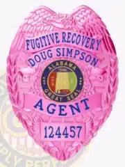 Pink BCAM Fugitive Recovery Agent badge with an eagle perched atop, displaying the name "Doug Simpson" in blue lettering, central Alabama Great Seal surrounded by a laurel wreath, a unique ID number 124457, and a safety pin fastener on the back.
