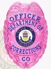 Detailed photo of a bright pink law enforcement badge for the Department of Corrections, showing an eagle crest on top, pink banners with blue lettering for "Officer Department of Corrections CO," the Colorado state emblem in the center, and a safety pin on the back.