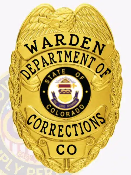 Custom gold metal badge with an eagle at the top, featuring black lettering for the Warden of the Department of Corrections, Colorado. The badge's center displays the official state seal, and it has a safety pin attachment.