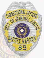 Detailed photo of a silver and gold law enforcement badge for the Department of Criminal Justice, showing an eagle crest on top, gold banners with blue lettering for "Correctional Officer Dept. of Criminal Justice Deputy Warden," the Texas state emblem in the center, and badge number 85.
