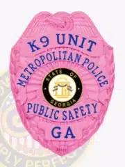 Detailed view of a pink eagle-shaped metal badge for Metropolitan Police K9 Unit, featuring blue text including "K9 UNIT," "METROPOLITAN POLICE," "PUBLIC SAFETY," and "GA," with the State of Georgia seal in the center and a safety pin attachment.