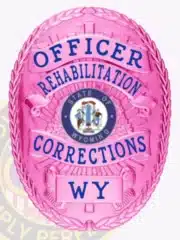 Pink corrections badge designed for an Officer in Wyoming Rehabilitation Corrections, with bold blue text on an oval-shaped BCAM pink background, detailed state seal in the middle, and a safety pin fastener.