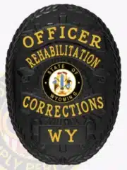 Oval-shaped black metal badge with gold letters reading "Officer Rehabilitation Corrections WY." The center displays the official Wyoming state seal, and the badge is equipped with a safety pin for secure attachment.